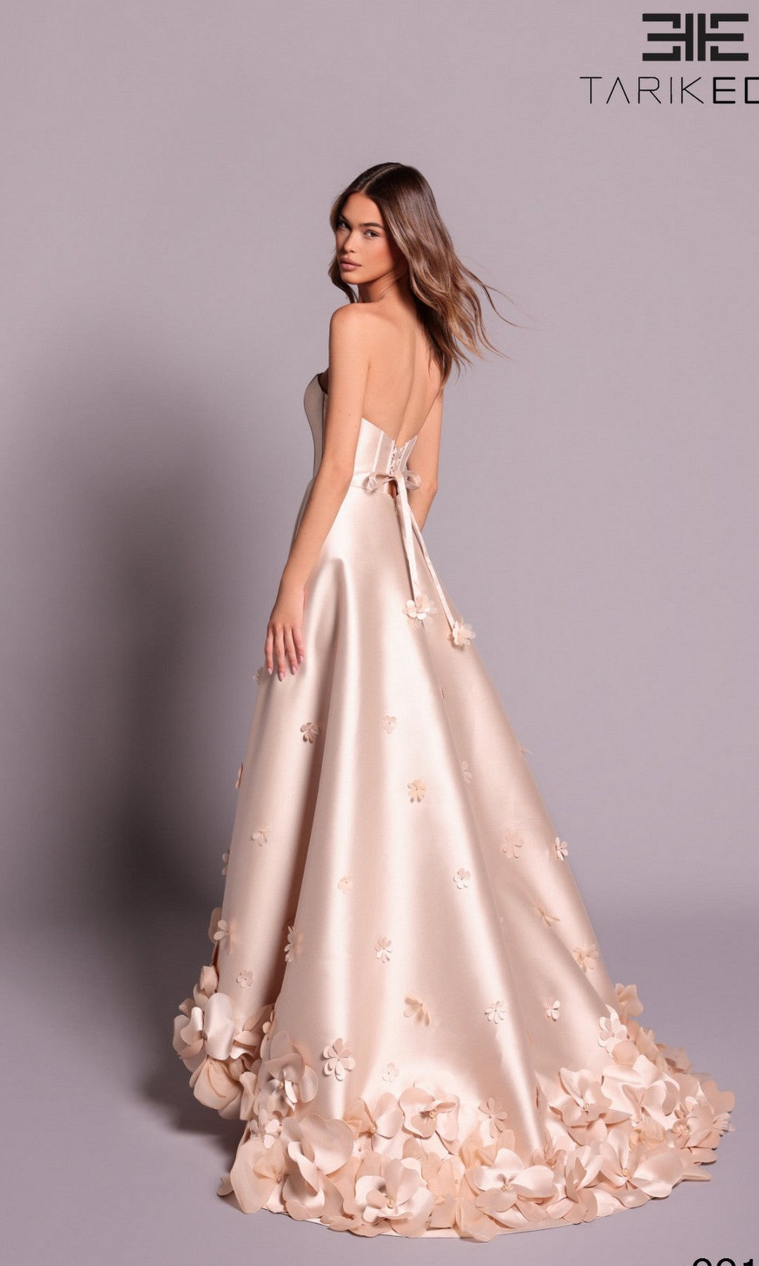 A woman stands against a plain background wearing the Tarik Ediz 99146: a champagne, strapless, floor-length formal dress with 3D flower appliqués and a flared hem.